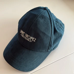 Grön keps Saint Tropez 1980 France - Snygg blå keps med böjd skärm och vit broderad text 'Saint Tropez 1980 France' framtill. Klassisk modell med paneler och knapp på toppen. Tillverkad i mjuk bomull som känns skön mot huvudet. Perfekt för dig som vill ha en chill och stilren look.