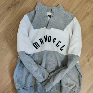 Grå och vit tröja från Manofell - Säljer en snygg grå och vit tröja från Manofell med tryckt logga över bröstet. Tröjan har lång ärm, ribbade muddar och knappar vid halsen. Perfekt för chill dagar och enkel att matcha med jeans eller joggers.