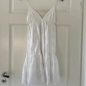 Vit kort sommarklänning med spets - Söt vit klänning med tunna axelband och v-ringad hals. Klänningen är kort och har broderade spetsdetaljer samt en volang nedtill. Perfekt för varma dagar, endast använd en gång 