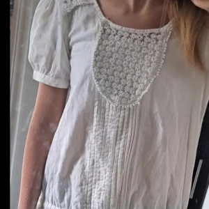 Vit blus - Supersöt vit blus med korta puffärmar. broderat blommönster vid halsen. Blusen har knytband i midjan och volang längst ner. Använd men absolut inga tecken på användning, bara lite svart på blommorna(bild finns). Står ingen storlek men skulle säga S, men man kan justera i midjan till tajtare.