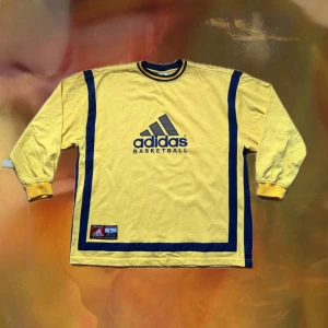 Gul Adidas baskettröja med blå detaljer - Retro gul långärmad baskettröja från Adidas med blå ränder längs sidorna och runt halsen. Stor Adidas-logga och 'BASKETBALL' tryckt på bröstet. Tröjan har ribbade muddar och är gjord i mjuk bomull. Perfekt för dig som gillar sportig och unik stil.