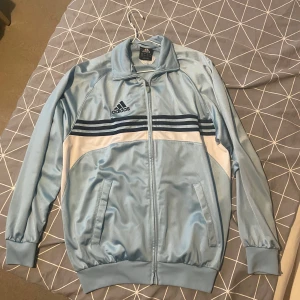 Ljusblå Adidas zip up - Snygg ljusblå Adidas tjocktröja med klassiska mörkblå ränder över bröstet och broderad logga. Passar även mig som är mer åt M hållet 