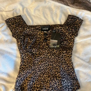 Leopardmönstrad sammetstopp Collectif - Supersnygg topp från Collectif i leopardmönstrad sammet med offshoulder-ärmar och knappar framtill. Modellen är figurnära och har dragkedja i sidan för enkel påklädning. Perfekt för dig som vill sticka ut med en djurisk look.