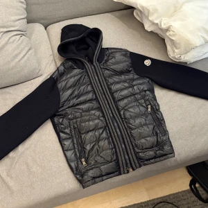 Moncler Cardigan - Säljer en svart cardigan jacka från Moncler i storlek L men passar L/M, quiltad front i syntet och stickade ärmar och huva. Jackan har dragkedja framtill, två fickor med dragkedja och Moncler-logga på ärmen. Perfekt för dig som vill ha en snygg och sportig look. Äktighetsbevis på bilderna. Priset är inte hugget i sten👍🏼. 
