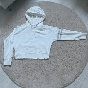 Vit sport hoodie från Stronger  - Säljer en fräsch vit hoodie från Stronger med två gråa ränder på ena ärmen. Tröjan har huva med snörning, ribbade muddar och är tillverkad i mjuk bomull. Perfekt för chill eller träning, riktigt clean look och skön passform.