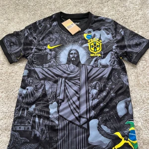 Svart Brasilien fotbollströja Nike - Unik svart Brasilien fotbollströja från Nike med detaljerat grafiskt tryck av Kristusstatyn och brasilianska symboler. Gula loggor och inslag av blått och grönt. Kortärmad modell i lätt, ventilerande material. Perfekt för dig som vill sticka ut på planen eller läktaren.