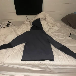 Nike svart och grå hoodiejacka - Nike hoodiejacka i svart och mörkgrått med hel dragkedja framtill och huva. Jackan har lång ärm, sportig look och är tillverkad i mjukt syntetmaterial. Perfekt för dig som gillar streetwear och vill ha en stilren jacka med tydlig Nike-logga på bröstet. 9/10 skick 