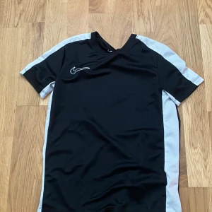 Svart Nike Dri-Fit fotbollströja M - Nike Dri-Fit fotbollströja i svart med vita paneler längs sidorna och axlarna. Tröjan har korta ärmar och rund hals. Materialet är lätt och ventilerande polyester, perfekt för träning eller match. Swoosh-logga broderad på bröstet.