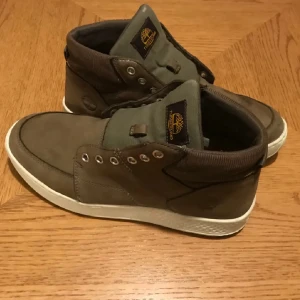 Bruna Timberland sneakers med vit sula - Snygga sneakers från Timberland i en brun och olivgrön färg med vit platt sula. Skorna har rund tå, snörning och detaljer i både skinn och textil. Märkeslogga på plösen och metallöglor för snören. Perfekt för dig som gillar street och outdoor-vibe.