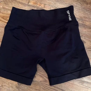 Svarta scrunch shorts från DFYNE - Svarta träningstights-shorts från DFYNE med scrunch-effekt bak för extra shaping. Högmidjade med bred ribbad linning som sitter skönt och ger stöd. Tight passform och stretchigt syntetmaterial som följer kroppen perfekt under gympasset.