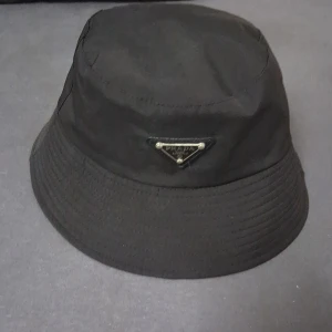 Svart bucket hat från Prada - Bucket hat i svart från Prada med klassisk rund form och bred brätte. Hatten har en ikonisk metalltriangel med Prada-logga framtill. Tillverkad i slitstark polyester som ger en clean och stilren look. Perfekt för dig som vill ha en cool accessoar med designer-vibe.
