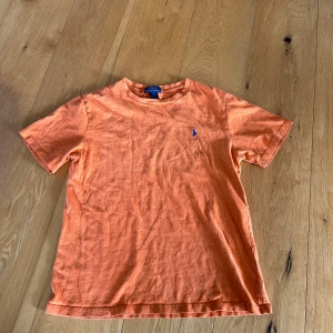Orange t-shirt från Polo Ralph Lauren - Snygg orange t-shirt från Polo Ralph Lauren med klassisk rund hals och korta ärmar. T-shirten har en liten blå broderad logga på bröstet och är gjord i mjuk bomull. Perfekt för dig som gillar stilrena och färgglada plagg.