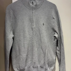 Grå half zip tröja Polo Ralph Lauren - Snygg grå långärmad tröja från Polo Ralph Lauren med half zip och diskret broderad logga på bröstet. Tröjan är stickad i bomull och har en klassisk, stilren look som passar perfekt till jeans eller chinos.