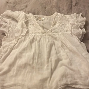 Vit blus med volang från Gina Tricot - Superfin vit blus från Gina Tricot med volangiga korta ärmar och v-ringning. Blusen har broderade detaljer upptill och är gjord i ett lätt, luftigt tyg som känns perfekt för varmare dagar. Snyggt flowy passform och romantisk vibe.