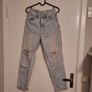 Ljusblå ripped jeans med hög midja - Säljer ett par ljusblå jeans med snygga slitningar på båda knäna. Jeansen har hög midja, raka ben och klassisk femficksdesign. Perfekta för en avslappnad och trendig look. Materialet är jeans i bomull.