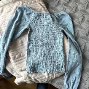 Ljusblå blus med puffärm - Supersöt ljusblå blus med smockad kropp och långa puffärmar i broderad bomull. Blusen har en offshoulder-design och är perfekt för vår och sommar. Materialet är mjukt och har fina hålbroderier på ärmarna som ger en romantisk vibe.