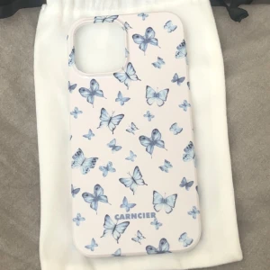 Telefonskal till iPhone 12 Pro Max - Säljer ett snyggt mobilskal från Carncier till iPhone 12 PRO Max med ett jätte fint fjärilsmönster i blått på en kräm vit bakgrund. Perfekt för att ge din telefon ett stilrent och unikt utseende.  Aldrig använt och nypris är 299kr