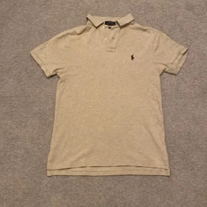 Beige kortärmad pikétröja från Ralph Lauren - Säljer en stilren beige pikétröja från Ralph Lauren i slim fit. Tröjan har korta ärmar och en klassisk krage med knappar. Den ikoniska broderade loggan syns på bröstet. Perfekt för en avslappnad men ändå elegant look.