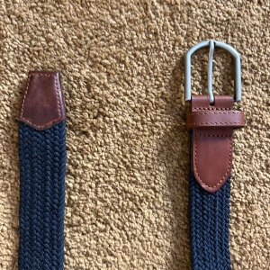 Mörk blått flätat bälte - Snyggt flätat bälte i mörkblått med bruna skinn detaljer och ett silverfärgat spänne. Perfekt för att ge en stilren touch till din outfit. Passar bra till både jeans och chinos.