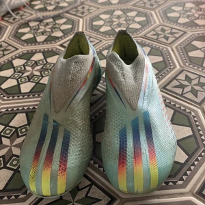 Flerfärgade fotbollsskor från Adidas - Snygga fotbollsskor från Adidas i en cool flerfärgad design med blå, gul och röd nyans. Skorna har en strömlinjeformad form utan snörning för en tight passform och optimal bollkontroll. Perfekta för fotbollsplanen!