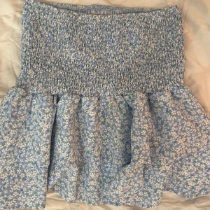 Blommig blå kjol - Söt blå kjol med blommigt mönster och smockdetaljer i midjan. Perfekt för en somrig look med sin luftiga design och fina volanger. 
