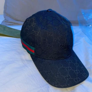 Svart keps från Gucci - Snygg svart keps från Gucci med diskret mönster och klassiska ränder i grönt och rött på sidan. Perfekt för att ge din outfit en lyxig touch. Kepsen har en justerbar rem för bästa passform.perfekt för sommaren.