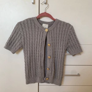stickad kortärmad kofta, grå - Säljer en stilren grå stickad kofta från H&M med korta ärmar och dekorativa guldknappar. Perfekt för lager-på-lager och ger en klassisk look. Passar bra till både jeans och kjol.