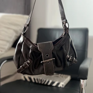 Brun axelväska med spännen - Snygg brun axelväska i skinn med dekorativa spännen och kontrastsömmar. Väskan har en justerbar axelrem och en praktisk dragkedja för säker förvaring. Perfekt för att ge din outfit en stilfull touch. 