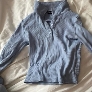 Ljusblå ribbad tröja från FB Sister - Säljer en ljusblå ribbad tröja från FB Sister i storlek XS. Tröjan har en hög krage och knappar framtill. Perfekt för en stilren och bekväm look.
