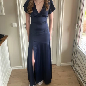 Mörkblå balklänning med slits - Elegant mörkblå långklänning med v-ringning och korta ärmar. Klänningen har en stilren slits framtill och är perfekt som balklänning. Materialet ger en fin glans och faller vackert. Pris går att diskutera🩷prislapp kvar, helt oanvänd💛