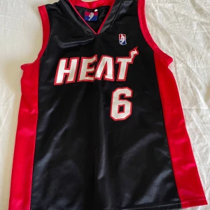 Svart och röd basketlinne - Säljer ett svart och rött basketlinne med texten 'HEAT' och nummer 6 på framsidan. Baksidan har namnet 'JAMES' och nummer 6. Perfekt för basketfans!