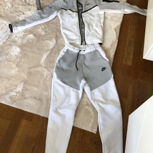 Nike teech fleece - Säljer en stilren vit och grå träningsdräkt från Nike. Jackan har dragkedja och svarta detaljer, medan byxorna har en elastisk midja och snörning. Perfekt för träning eller en avslappnad dag.