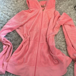 Rosa hoodie från Juicy Couture - Säljer en mjuk och gosig rosa hoodie från Juicy Couture. Den har en dragkedja framtill och en stor luva. Baksidan pryds av texten 'Juicy' i svart med dekorativa hjärtan runtom. Perfekt för en avslappnad stil!