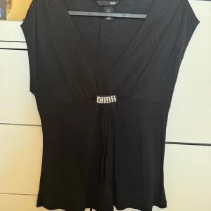 Svart topp från H&M med dekorativ detalj - Snygg svart topp från H&M med en v-ringad design och en glittrig detalj framtill. Toppen har korta ärmar och en elegant passform som passar perfekt för en kväll ute.