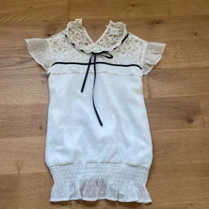 Vacker vit blus med spetsdetaljer från Lindex - Säljer en söt vit blus från Lindex med spetsdetaljer och en liten rosett vid halsen. Blusen har korta ärmar och en volangkant . Perfekt till sommaren