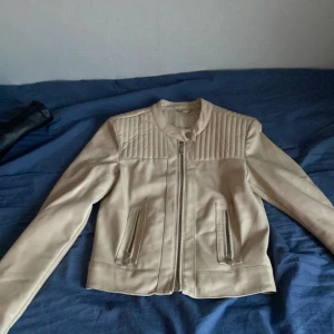 Beige skinnjacka från Amisu - Säljer en stilren beige skinnjacka från Amisu. Jackan har en dragkedja framtill och quiltade detaljer på axlarna. Perfekt för en trendig look!