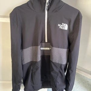 Svart vindjacka från The North Face - Säljer en svart vindjacka från The North Face med vit dragkedja och logga på bröstet. Jackan har en huva och är tillverkad i ett lätt syntetmaterial, perfekt för blåsiga dagar. Den har långa ärmar och en praktisk design.