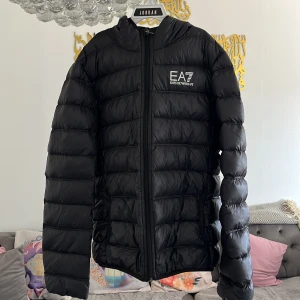 Svart dunjacka från EA7 Emporio Armani - Snygg svart dunjacka från EA7 Emporio Armani med dragkedja och huva. Jackan har en quiltad design och är perfekt för kyligare väder. Den har en normal passform och är långärmad.