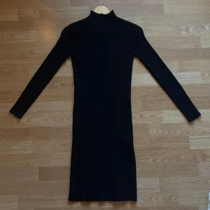 Svart ribbad klänning - Snygg svart ribbad klänning med lång ärm och hög krage. Perfekt för en stilren look. Klänningen har en figurnära passform och är gjord i ett stretchigt material.