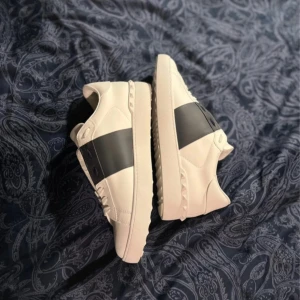 Valentino Open sneakers i vitt och svart - Snygga Valentino Open sneakers i vitt skinn med en bred svart rem över ovansidan. Skorna har en rund tå och klassisk snörning. Perfekta för en stilren look.