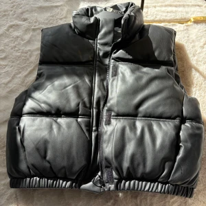 Svart väst coatad med dragkedja - Snygg svart dunväst med dragkedja och hög krage. Perfekt för kyliga dagar när du vill hålla värmen utan att kompromissa med stilen. Västen har en stilren design och passar till många olika outfits.