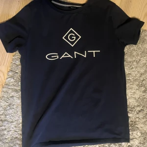Svart t-shirt från GANT - Snygg svart t-shirt från GANT med deras ikoniska logga tryckt på framsidan. Perfekt för en stilren och avslappnad look. Tillverkad i mjukt material för extra komfort. Väldigt bra skick utan defekter. 
