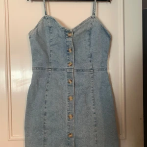 Jeansklänning med knappar - Trendig jeansklänning med smala axelband och knappar framtill. Perfekt för en avslappnad stil. Klänningen har en figurnära passform och är tillverkad i ett klassiskt denimtyg.