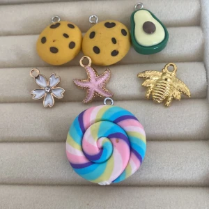 Charmiga berlocker i olika former - Säljer ett set med sju unika berlocker: en färgglad spiral, två kakor med chokladbitar, en avokado, en blomma med strass, en sjöstjärna och ett bi i guld. Perfekta för att göra dina smycken personliga och roliga!