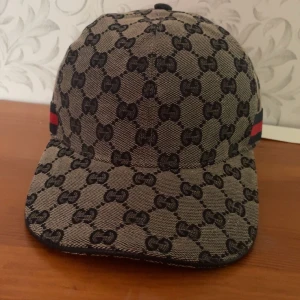 Beige mönstrad keps - Snygg beige keps med ikoniskt mönster och röd-blå randdetalj på sidan. Perfekt för att ge din outfit en lyxig touch.