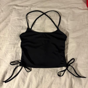 Svart ärmlös linnetopp - Snygg svart linnetopp med smala axelband som korsas i ryggen. Toppen har knytband i sidorna för justerbar passform och en stilren look. Perfekt för en trendig och avslappnad stil.