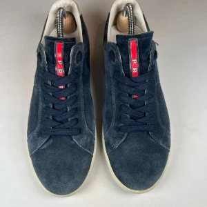 Mörkblå mockasneakers från Prada - Snygga mörkblå sneakers i mocka från Prada med vit sula och klassisk snörning. De har en stilren design med en röd detalj på plösen som ger en extra touch. Perfekta för en avslappnad men ändå stilfull look.