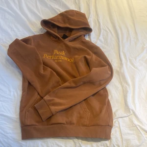 Brun hoodie från Peak Performance - Säljer en snygg brun hoodie från Peak Performance med broderad logga i gult på bröstet. Tröjan har en klassisk känguruficka och ribbade muddar vid ärmslut och nederkant. Perfekt för en avslappnad stil! Pris kan diskuteras