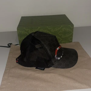 Svart keps med ormmotiv från Gucci - Snygg svart keps från Gucci med ett iögonfallande ormmotiv i rött och vitt. Kepsen har ett diskret mönster och justerbar rem baktill. Perfekt för att ge din outfit en lyxig touch.