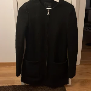 Svart kappa från New Look - Snygg svart kappa från New Look med dragkedja framtill. Den har en stilren design med långa ärmar och praktiska fickor. Perfekt för både höst och vår.